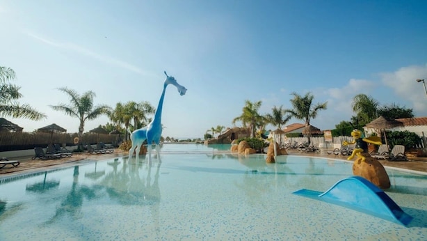 Zona de piscina con estatua de jirafa, sillas de descanso, palmeras y cielo despejado.