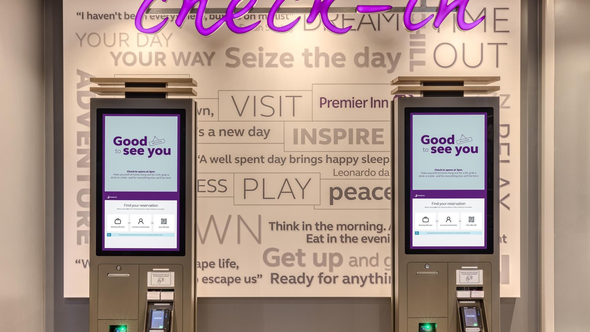 Deux bornes d’enregistrement dotées d’écrans affichant un message de bienvenue, un code QR permettant de retrouver les réservations, et un mur orné de phrases motivantes.