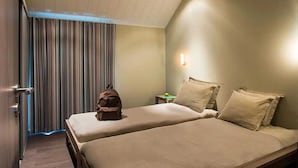 Quarto de hotel com duas camas, cortina listrada, mesa de cabeceira com abajur e uma mala em uma das camas.