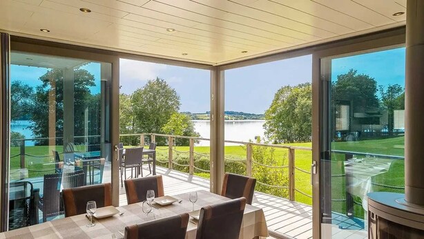 Uma área de refeições com uma mesa posta para dois, com vista para um lago panorâmico e paisagem gramada.