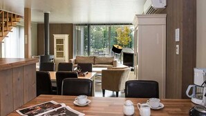 Uma sala de estar moderna com uma mesa de madeira, cadeiras, um sofá e uma televisão. Há também uma cozinha com um frigorífico e uma máquina de café.
