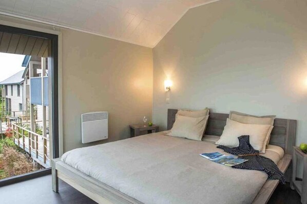 Une chambre avec un grand lit, une table de chevet, un radiateur fixé au mur et une fenêtre donnant sur une maison et de la verdure.