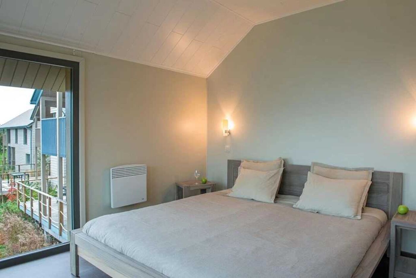 Ein Schlafzimmer mit einem großen Bett, einem Nachttisch, einer Wandleuchte und einem Fenster mit Blick auf einen Balkon.