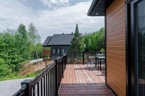 A wooden deck with a railing, a table, and chairs, overlooking a forested area. - Le point de rencontre des Éboulements (Les Éboulements)