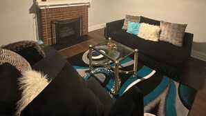 Un salon avec une cheminée, un canapé noir, une table basse en verre et un téléviseur qui affiche l’heure.