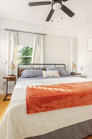 A bedroom with a bed, a ceiling fan, a window with curtains, and a bedside table with a lamp.