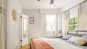 A bedroom with a bed, a bedside table, a window with curtains, and a door. - Lil’ Abbottry - 1/1 Apt - 5 Min Walk to City Gates (St. Augustine)