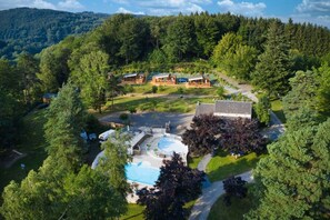 An aerial view of a resort with multiple cabins, a swimming pool, and a forested backdrop.