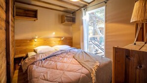 A cozy bedroom with a wooden headboard, a large bed with a pink quilt, a wooden nightstand, and a window with a view of outdoor equipment. - ZEcamping 3 étoiles pataugeoire piscine dog friendly locatif LV 40m2 (Le Bez)
