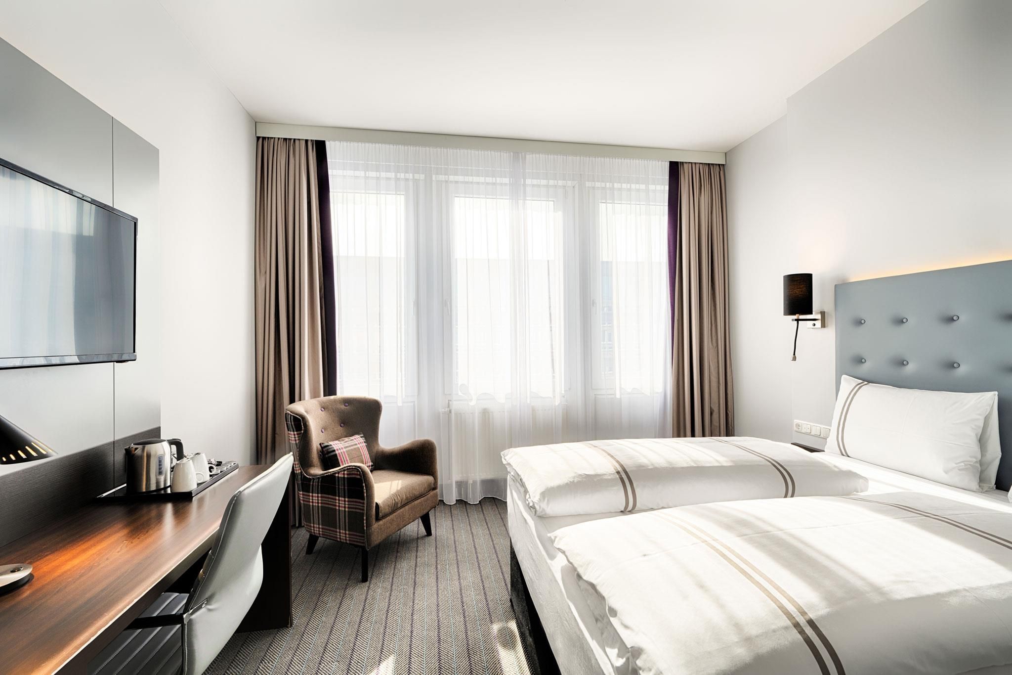 Habitación de hotel con cama, una silla, un escritorio con tetera, televisión y ventana con cortinas.