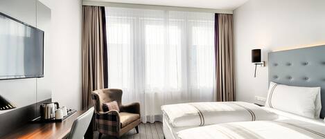 Habitación de hotel con cama, una silla, un escritorio con tetera, televisión y ventana con cortinas.