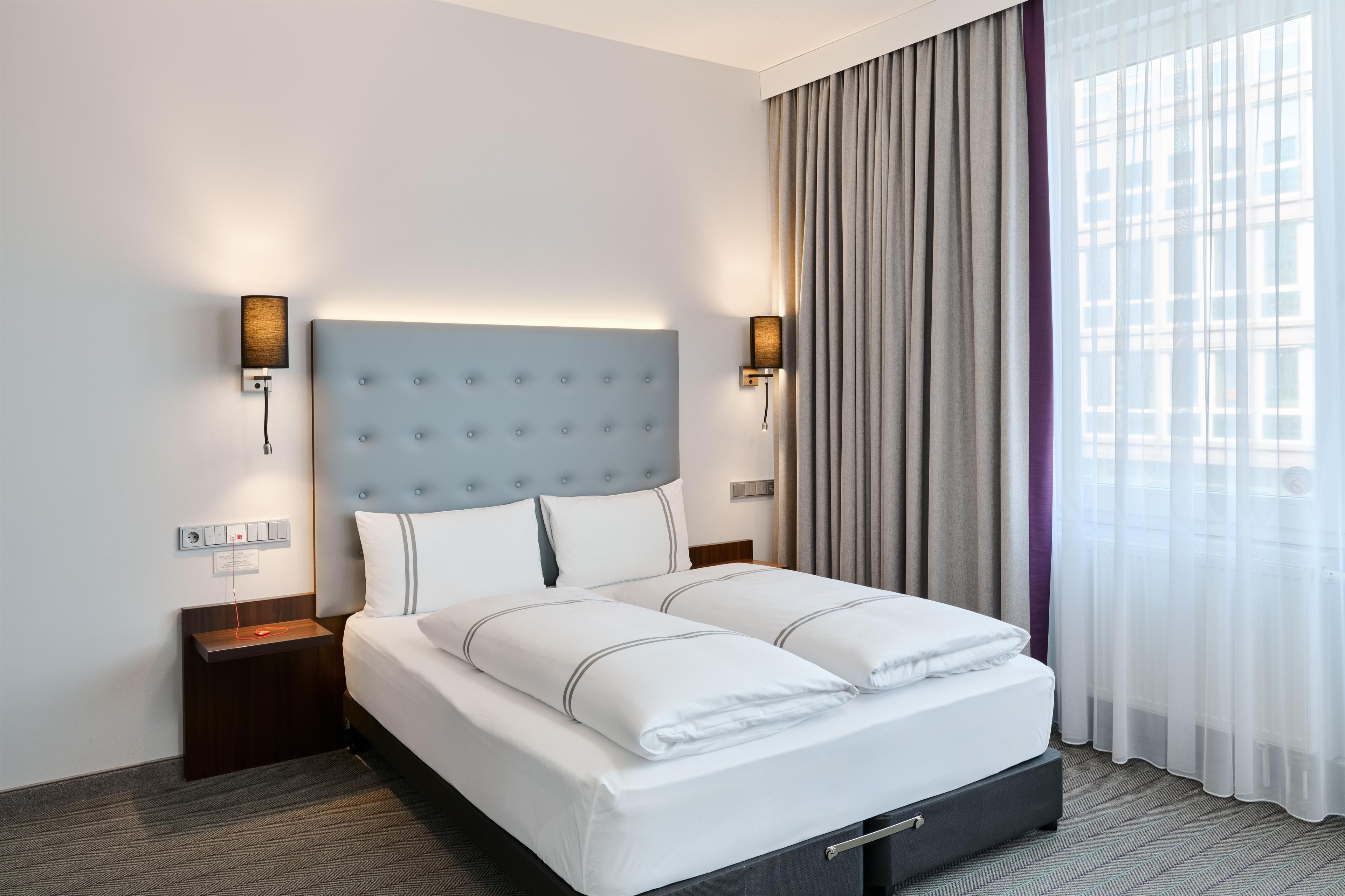 A hotel room with a large bed, two bedside tables, a wall-mounted lamp, and a window with curtains.