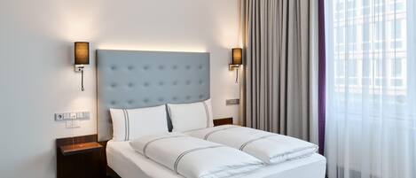 A hotel room with a large bed, two bedside tables, a wall-mounted lamp, and a window with curtains.