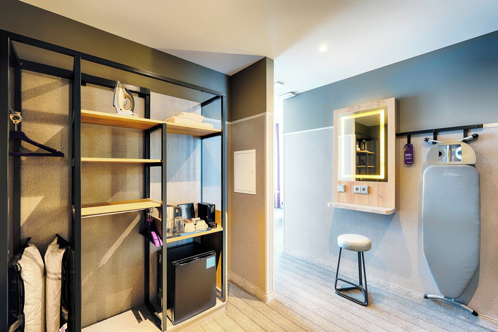Ein modernes Hotelzimmer mit begehbarem Kleiderschrank, einer kleinen Küchenzeile, einem Spiegel mit Beleuchtung und einem tragbaren Bügeleisen.