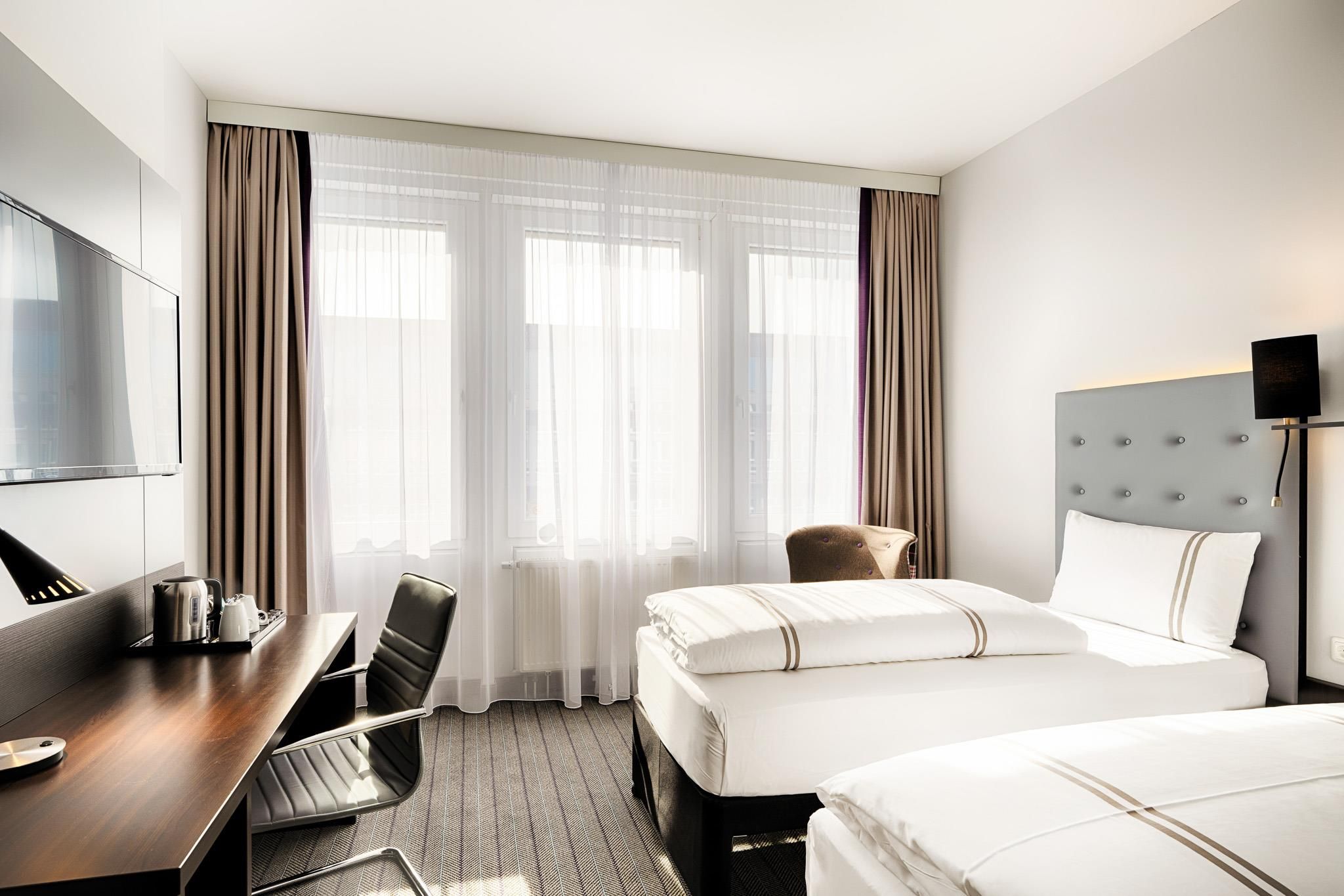 A hotel room with a bed, a desk with a chair, a television, and a window with curtains.