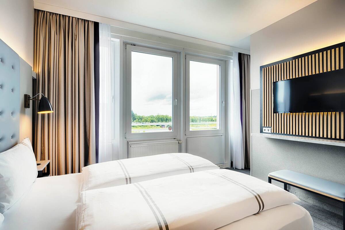 Habitación de hotel con una cama grande, un televisor montado en la pared y una ventana con cortinas.