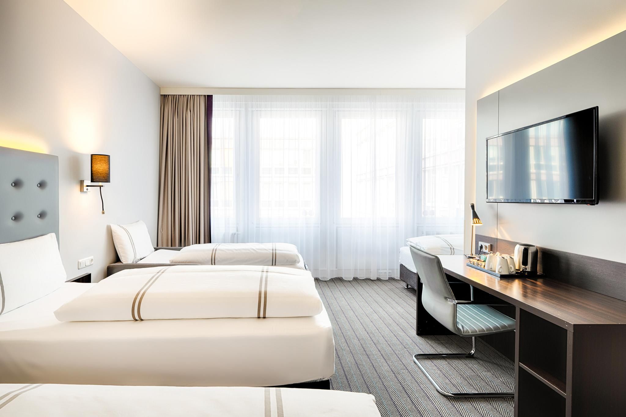 Ein Hotelzimmer mit zwei Betten, einem Schreibtisch, einem Stuhl, einem Flachbildfernseher und einem Fenster mit Vorhängen.
