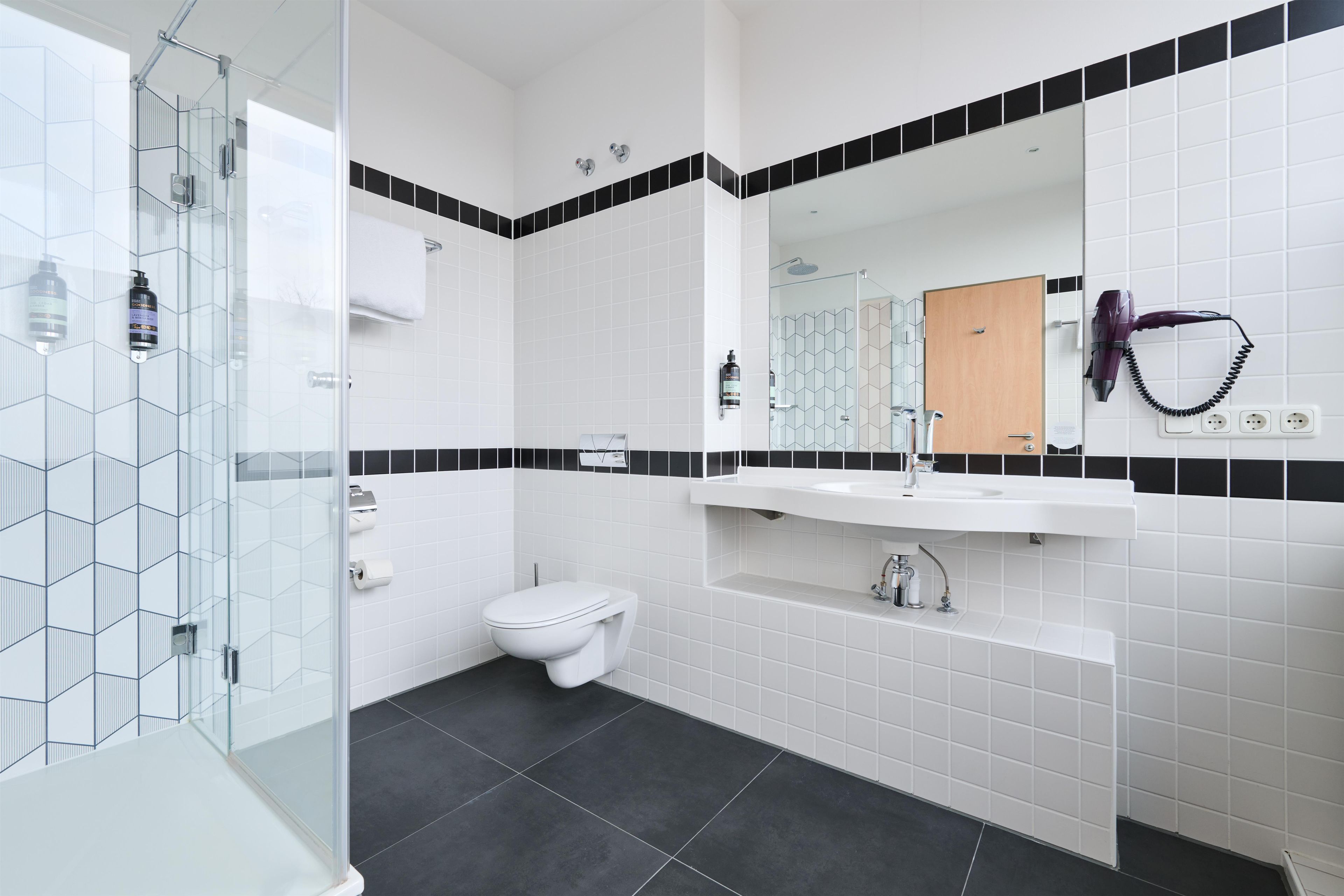 Een moderne badkamer met een douchecabine van glas, een wit toilet, een witte wastafel met een spiegel erboven en een vloer van zwart tegels.