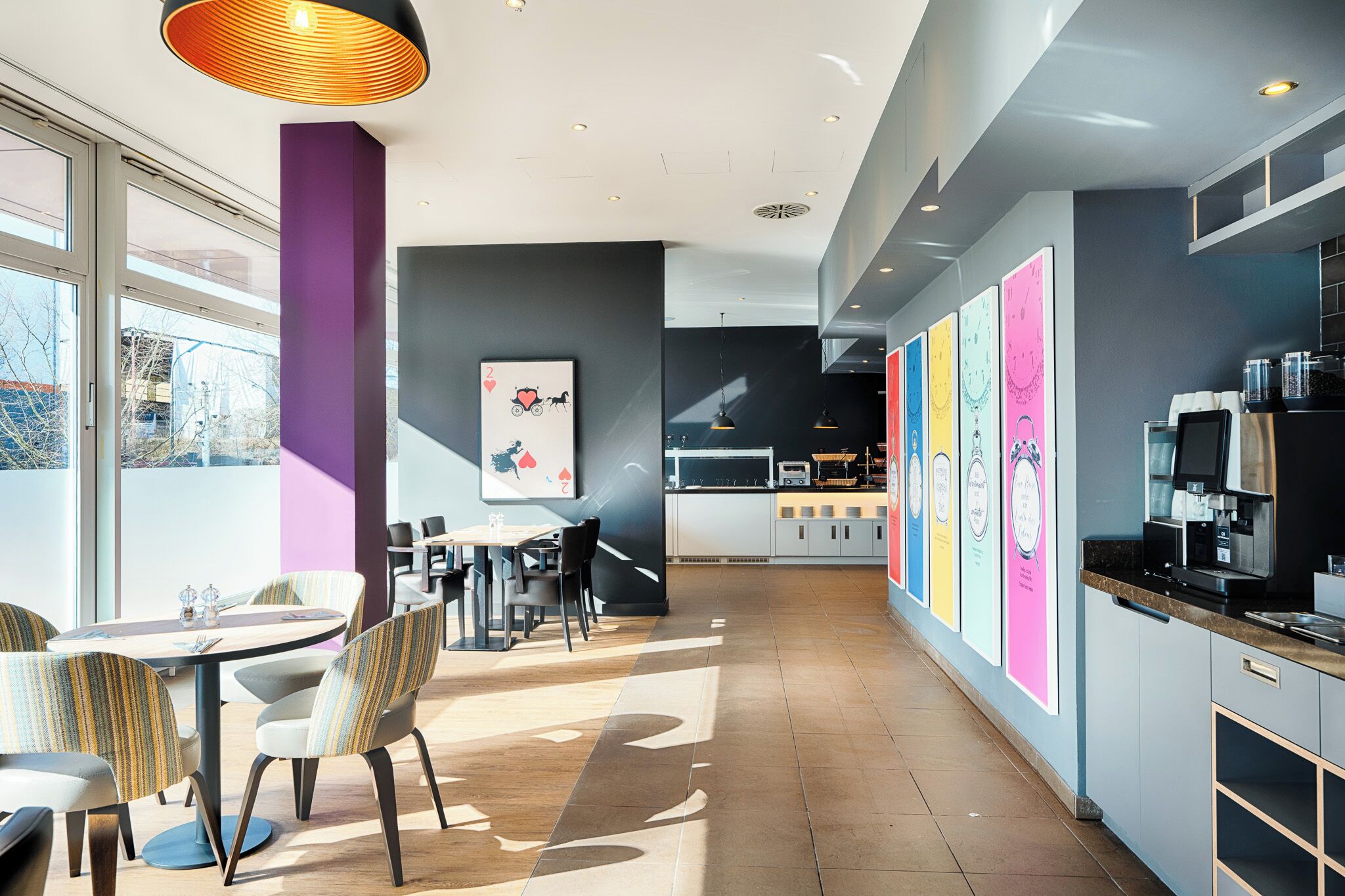A modern restaurant with a long counter, tables, and chairs. There are colorful posters on the wall and a large window providing natural light.
