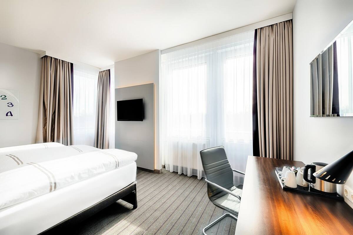 Una habitación de hotel con cama, escritorio con lámpara, silla, televisión y cortinas.