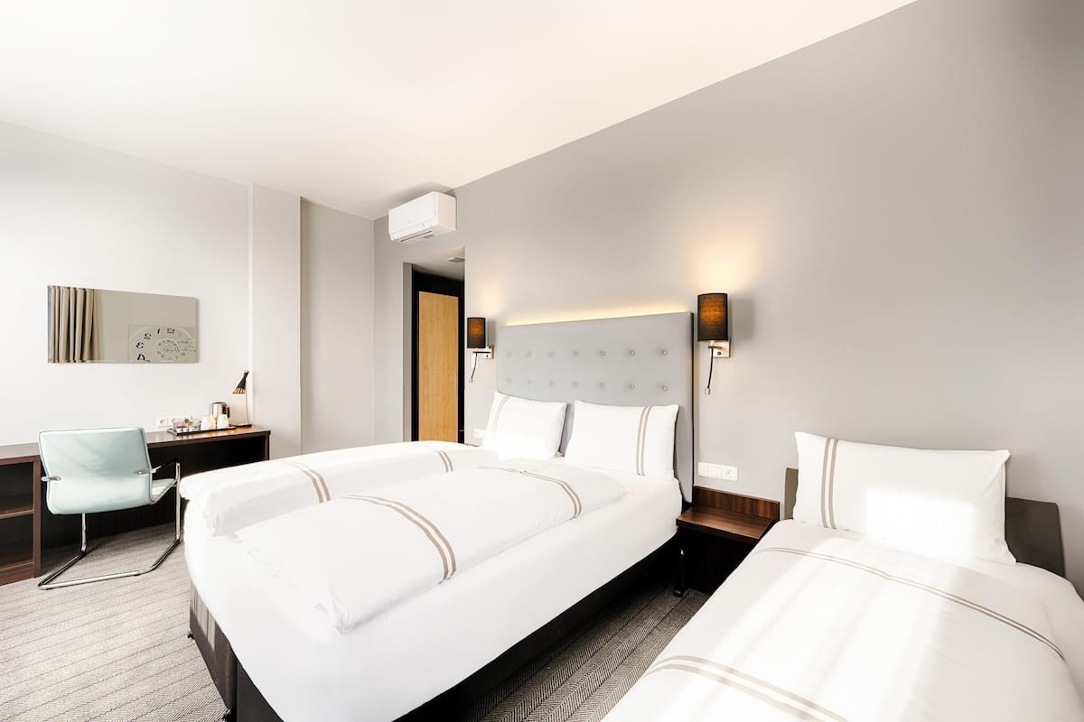 Quarto de hotel com duas camas, uma escrivaninha, uma cadeira, um relógio de parede e uma luminária de parede.