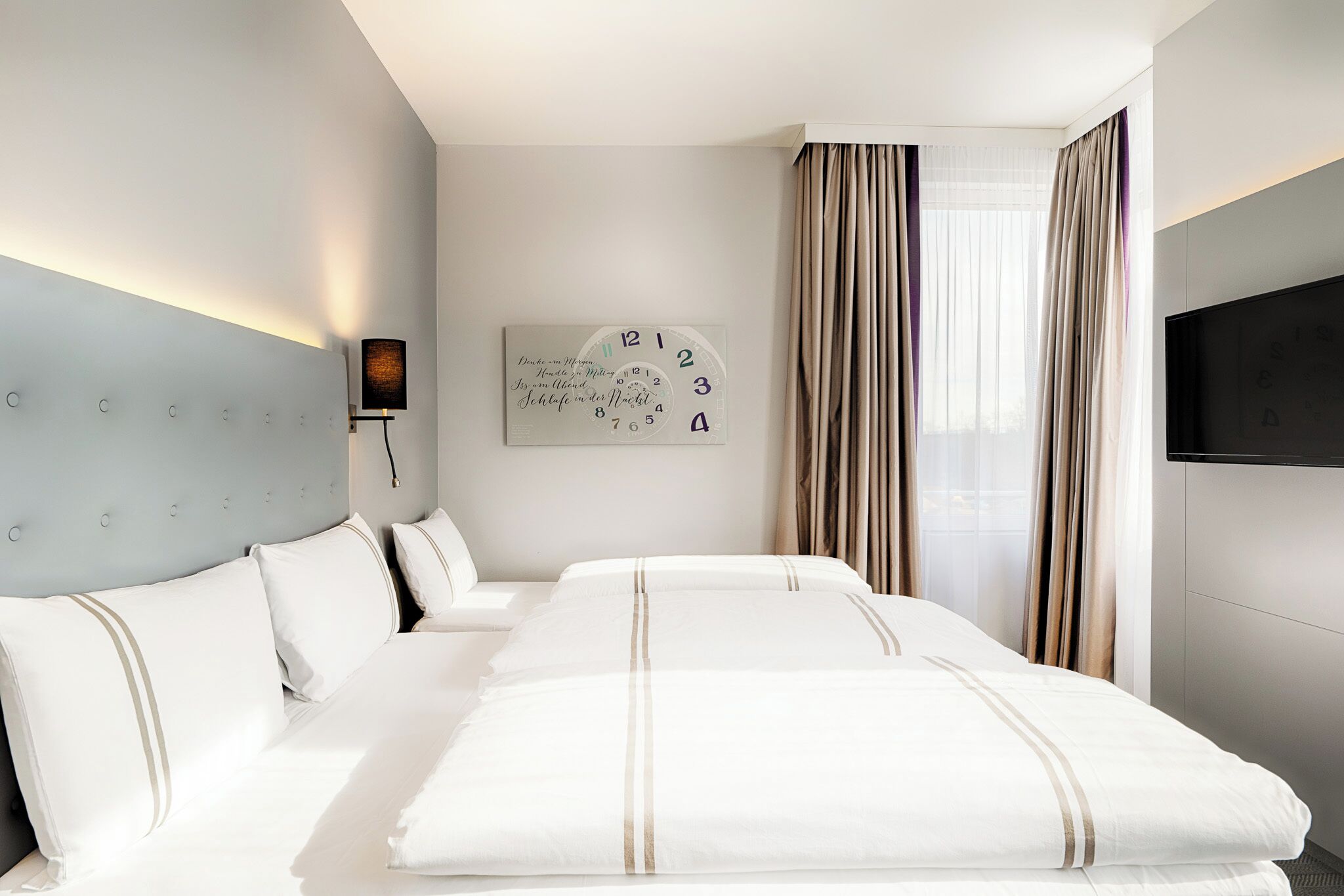 Habitación de hotel con una cama grande, un reloj de pared, un televisor de pantalla plana y una ventana con cortinas.