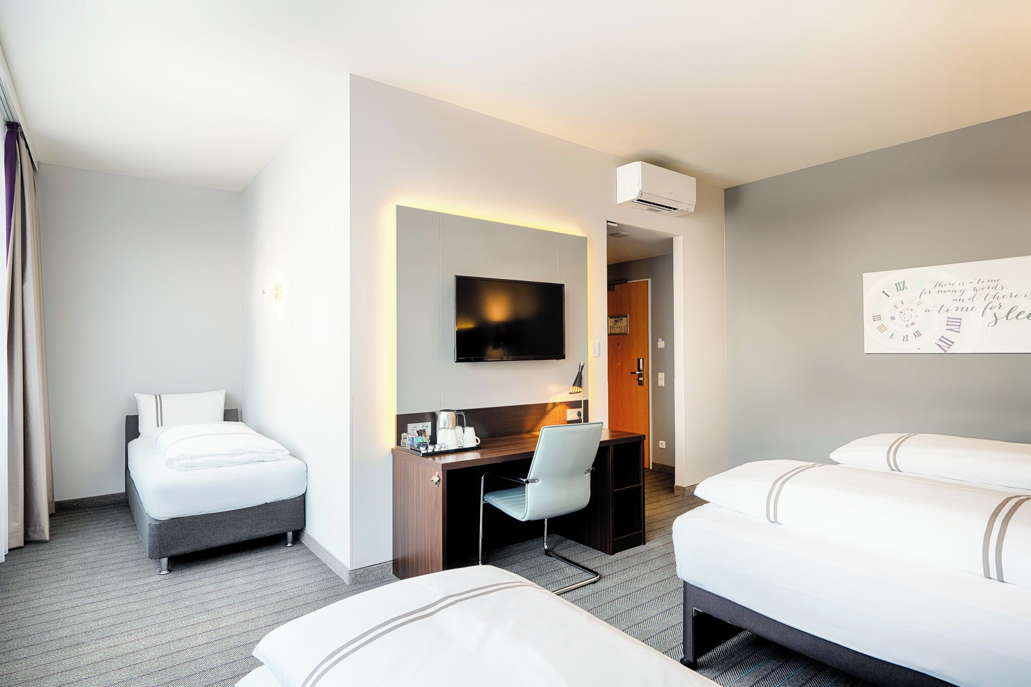 Habitación de hotel con dos camas, un escritorio con silla, televisor de pantalla plana y un reloj de pared.