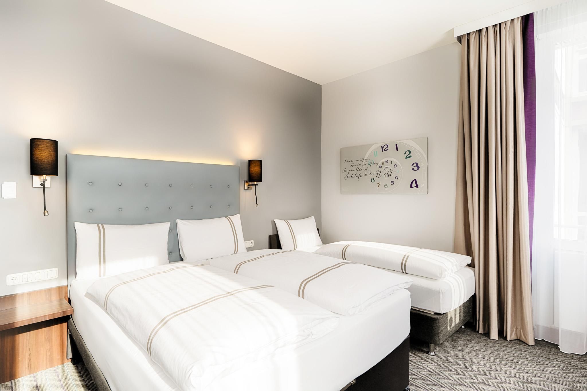 Habitación de hotel con una cama grande, dos mesitas de noche, un reloj de pared y una ventana con cortinas.