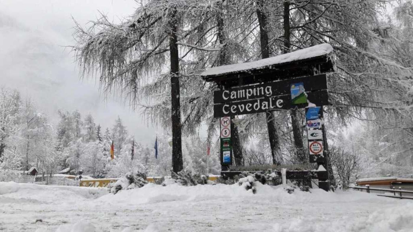 Un panneau indicateur recouvert de neige, portant l’inscription « Camping Cevedale » et divers symboles indiquant les équipements et les restrictions.