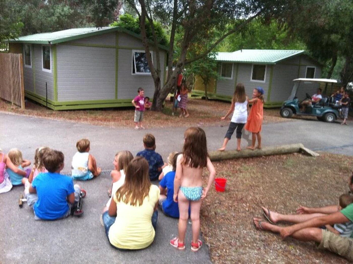 A group of children sitting on the ground in front of a small building, possibly a school or community center.