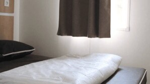 A single bed with a white mattress and a dark headboard, a window with brown curtains, and a wooden shelf with a picture frame.