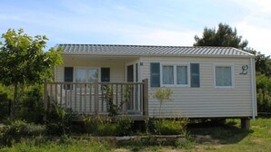 A single-story mobile home with a porch and a small deck, surrounded by greenery.
