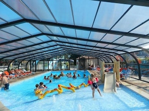 Piscina coberta com pessoas nadando e brincando, um escorregador e cadeiras de descanso.
