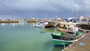 A marina with several boats docked, a lighthouse in the background, and a row of parked vehicles along the shore.