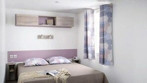 A bedroom with a bed, a nightstand, a wall-mounted shelf, and a window with curtains. - Camping 4 étoiles - Piscine  - efchaa (Bourgneuf-en-Retz)