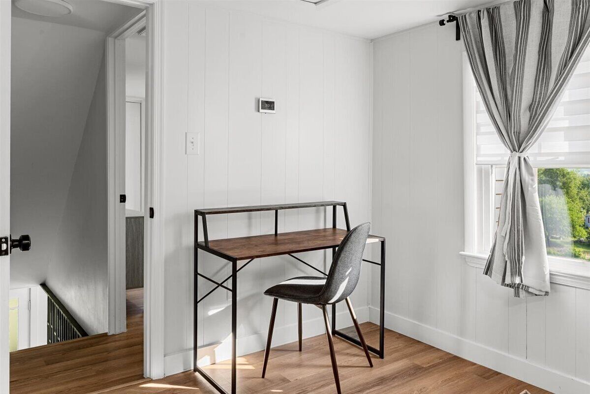 A room with a wooden desk and chair, white walls, and a window with curtains.
