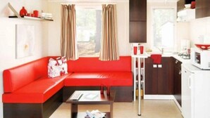 A modern living room with a red sofa, a coffee table, and a kitchen area with a microwave and cabinets. - ZEcamping 4* animé piscine couverte mobil-home TV BBQ 30m2 6 pers (Bourgneuf-en-Retz)