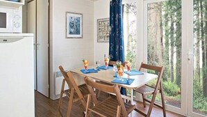 Ein Essbereich mit einem Tisch für vier Personen, Holzstühlen, einem weißen Kühlschrank und Blick auf Bäume durch eine Glastür.