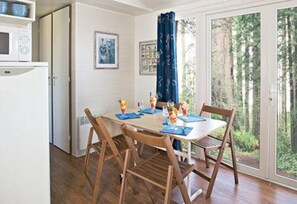 Un coin repas avec une table dressée pour quatre personnes, des chaises en bois, un réfrigérateur blanc et une vue sur les arbres à travers une porte vitrée.