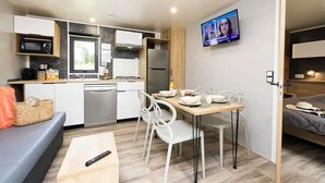 A compact kitchen with modern appliances, a dining area with white chairs, and a TV mounted on the wall. - ZEcamping 4 étoiles animé parc aquatique mobil-home TV LV BBQ 32m2 (Loupiac)