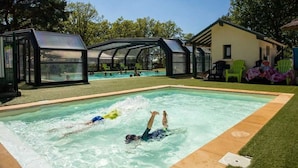 A swimming pool with two people swimming, surrounded by a covered area with chairs and a building. - ZEcamping 4 étoiles animé parc aquatique mobil-home TV LV BBQ 32m2 (Loupiac)