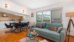 Een moderne woonkamer met een bank, een eethoek met een tafel en stoelen, een keuken met een fornuis, en een balkon met uitzicht op bergen.