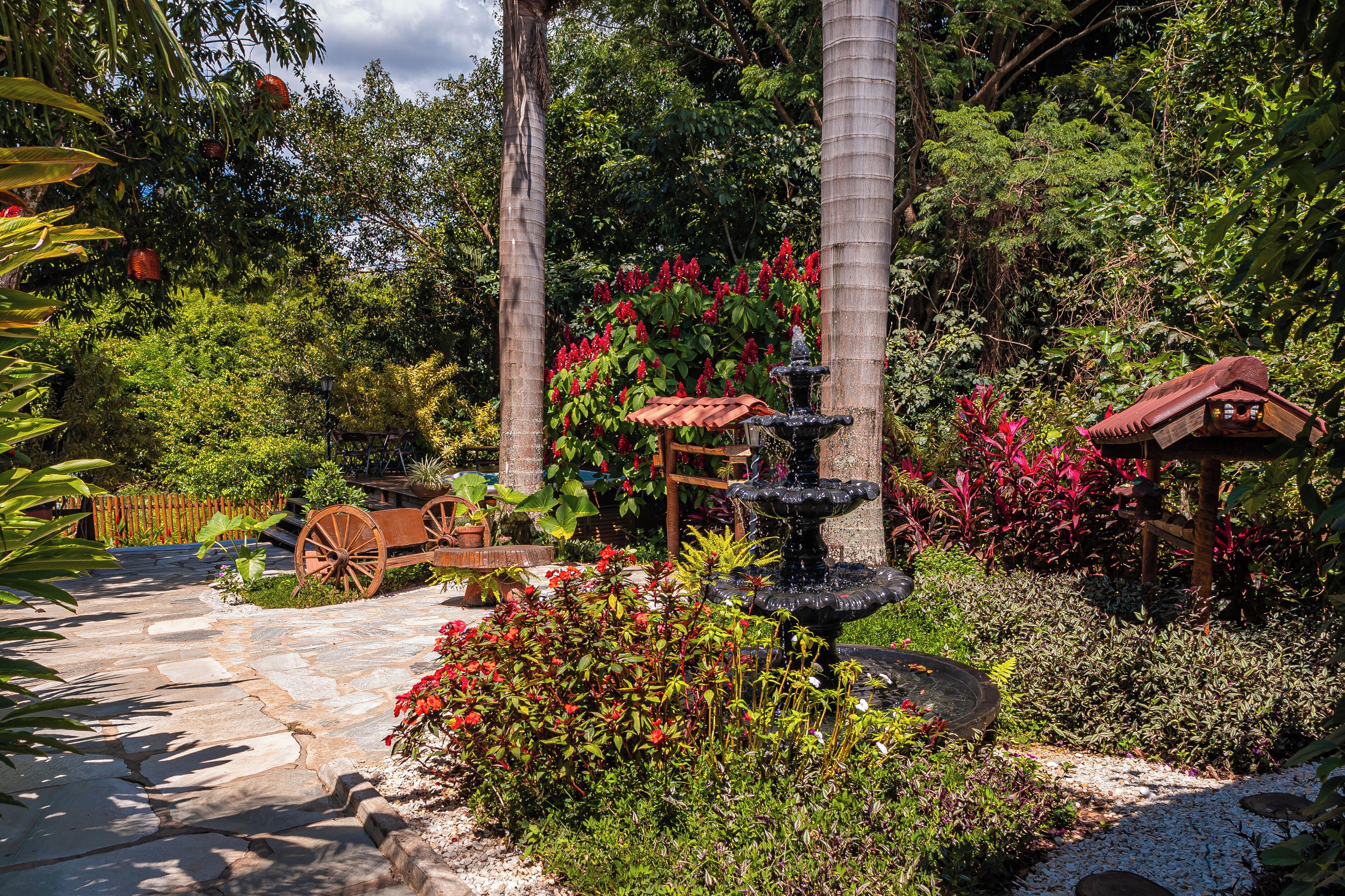 A garden with a stone pathway, a wooden cart, a fountain, and lush greenery.