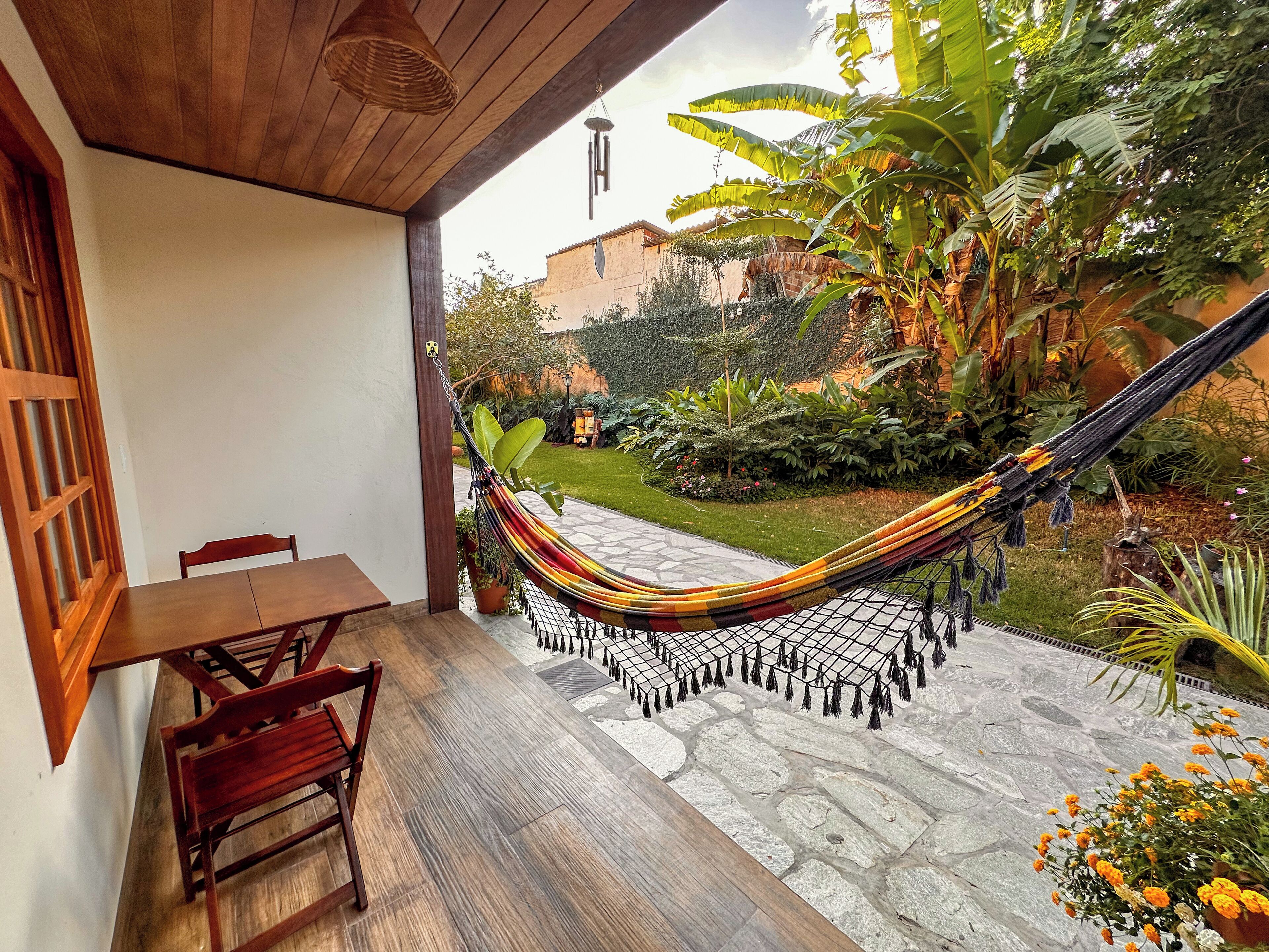 A hammock on a patio with a wooden table and chairs, surrounded by lush greenery and a stone pathway.
