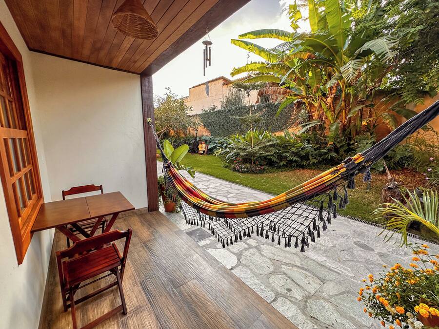 A hammock on a patio with a wooden table and chairs, surrounded by lush greenery and a stone pathway.