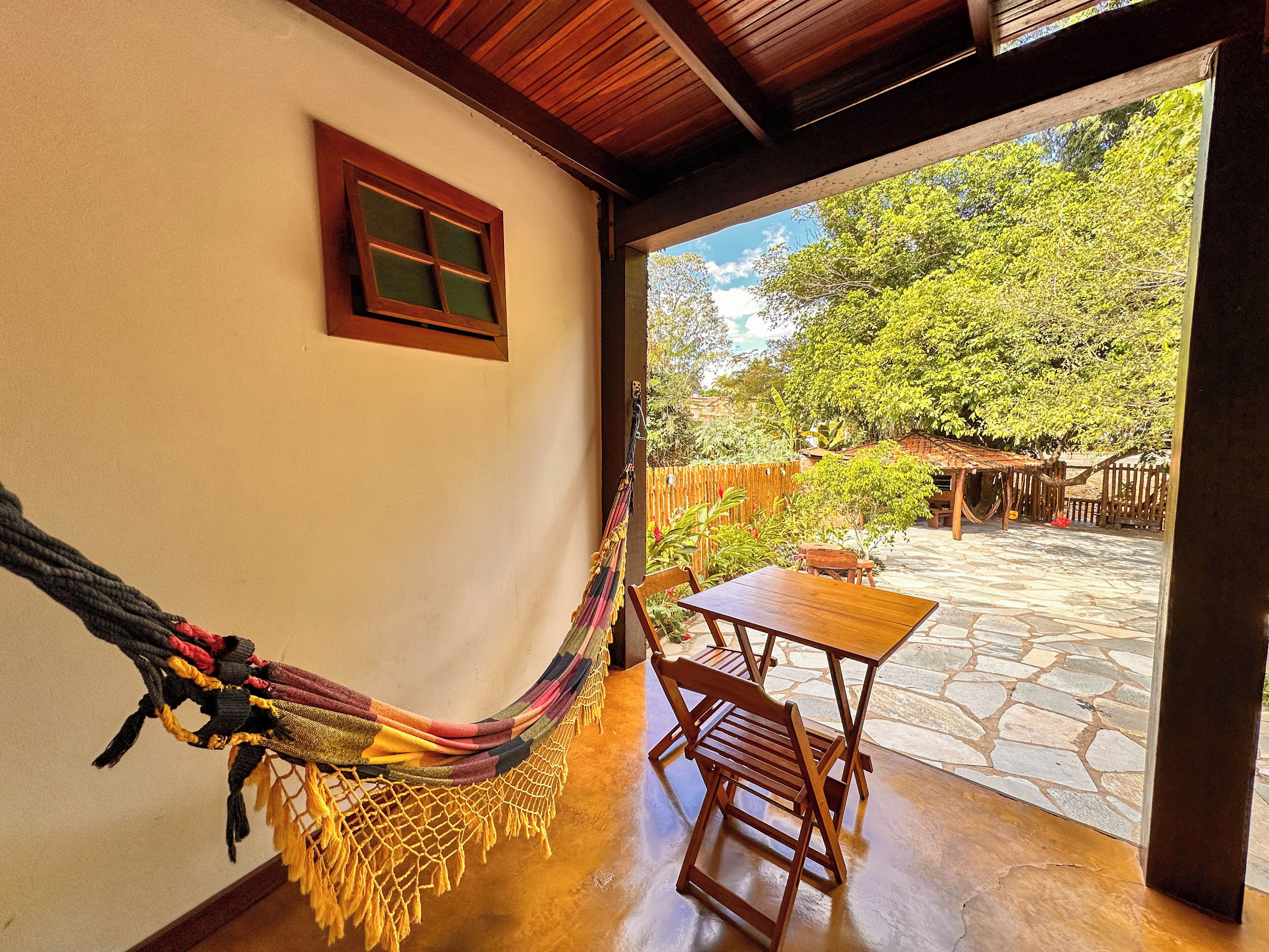 A hammock, wooden table, and chairs in a room with a stone floor and a view of greenery outside.