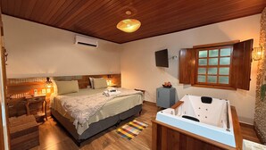 Un dormitorio con cama, mesitas de noche, un escritorio pequeño, una lámpara de escritorio, un televisor montado en la pared, una ventana con contraventanas de madera, una bañera empotrada y suelo de madera.