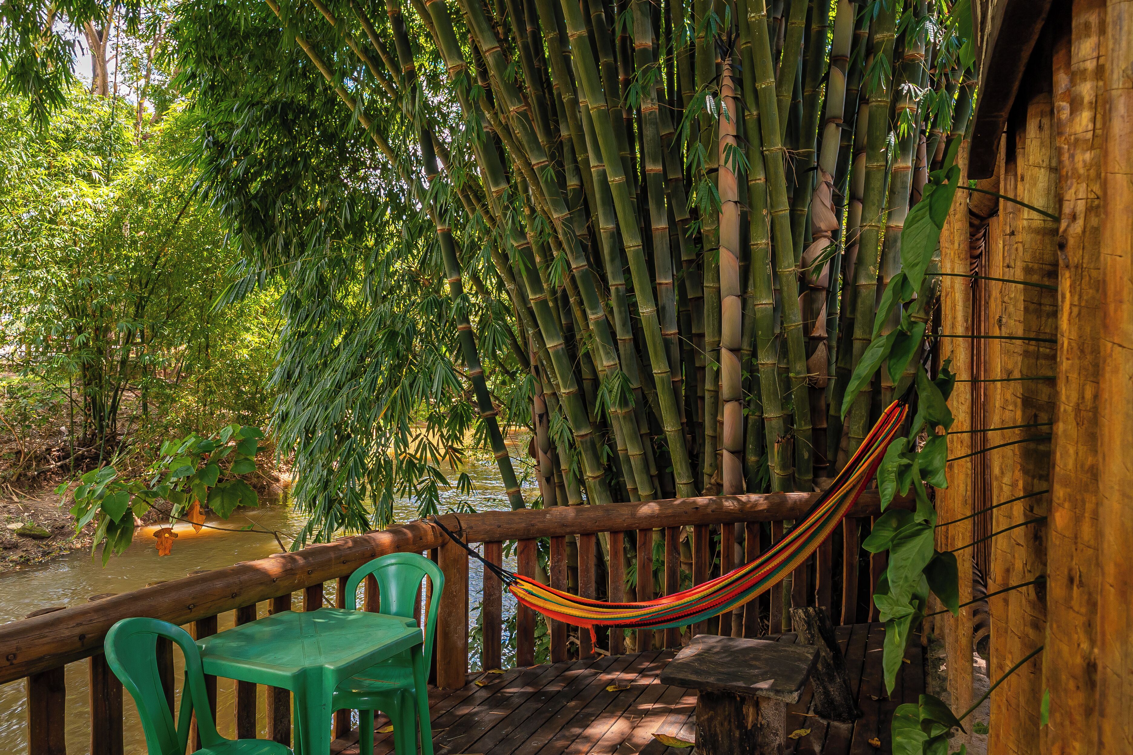 A wooden deck with a hammock, green plastic chairs, and a table overlooking a bamboo forest and a body of water.