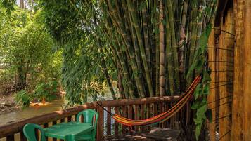 A wooden deck with a hammock, green plastic chairs, and a table overlooking a bamboo forest and a body of water.