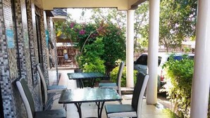An outdoor patio area with tiled flooring, a covered seating area with metal tables and chairs, and a view of greenery and parked cars in the background. - Acacia Shade Hotel and Gardens (Mutomo)